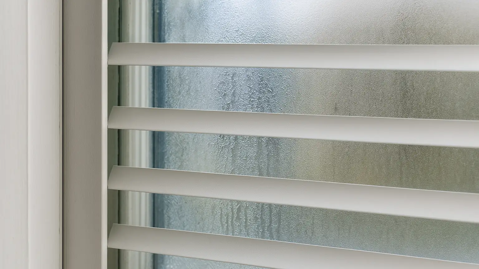 Close-up of a window pane with closed blinds directly against the glass, showing the narrow gap between slats and window surface, with visible heat distortion or condensation patterns on the glass center versus cooler edges.